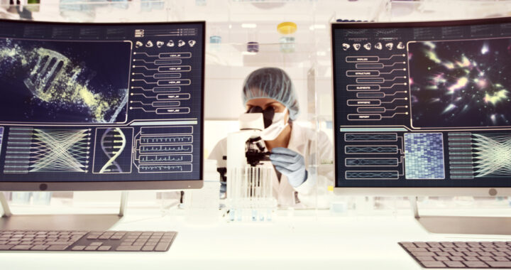 A scientist looks into a microscope next to monitors displaying DNA graphics