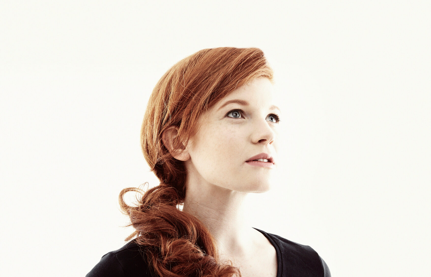A woman in her twenties with red hair looks thoughtfully toward the sky.