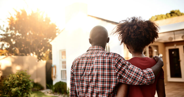 A man and a woman look at a house with their arms around each other.