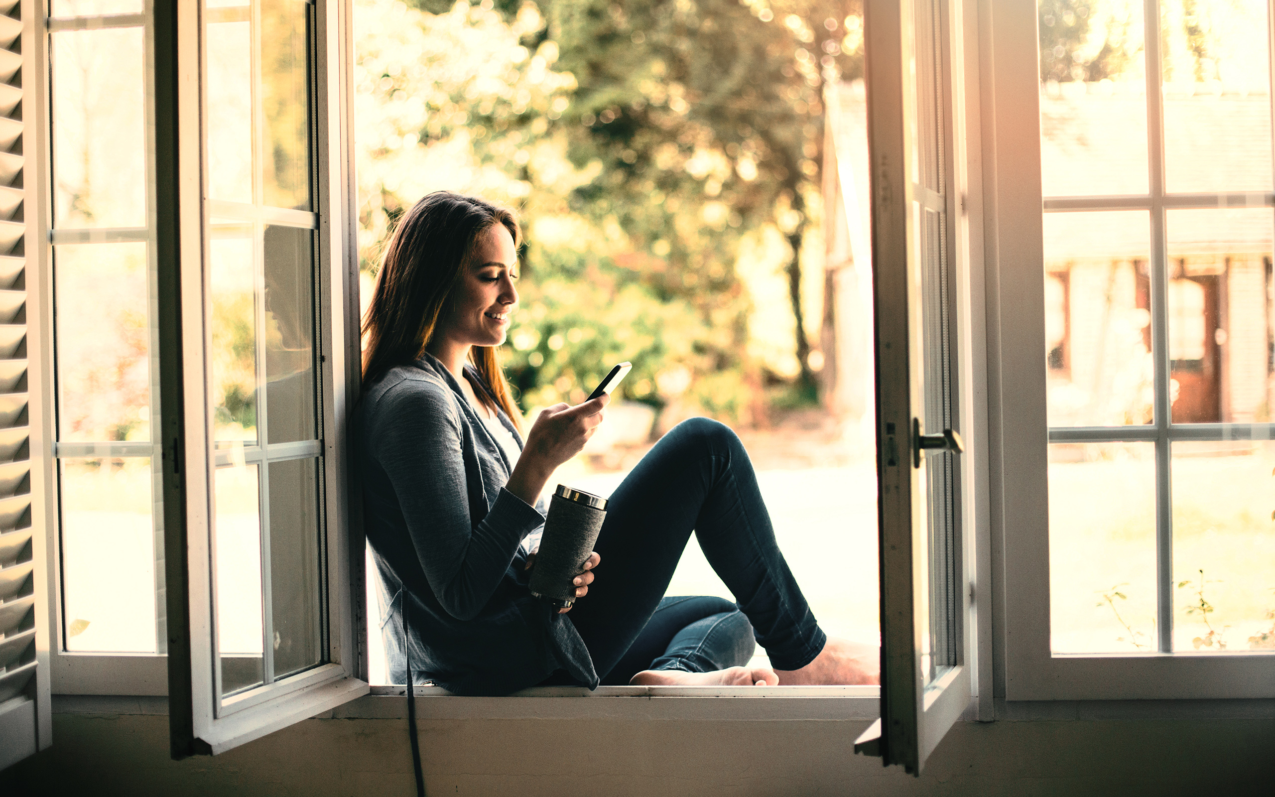 A woman sitting on the ledge of an open window, looking at her phone.