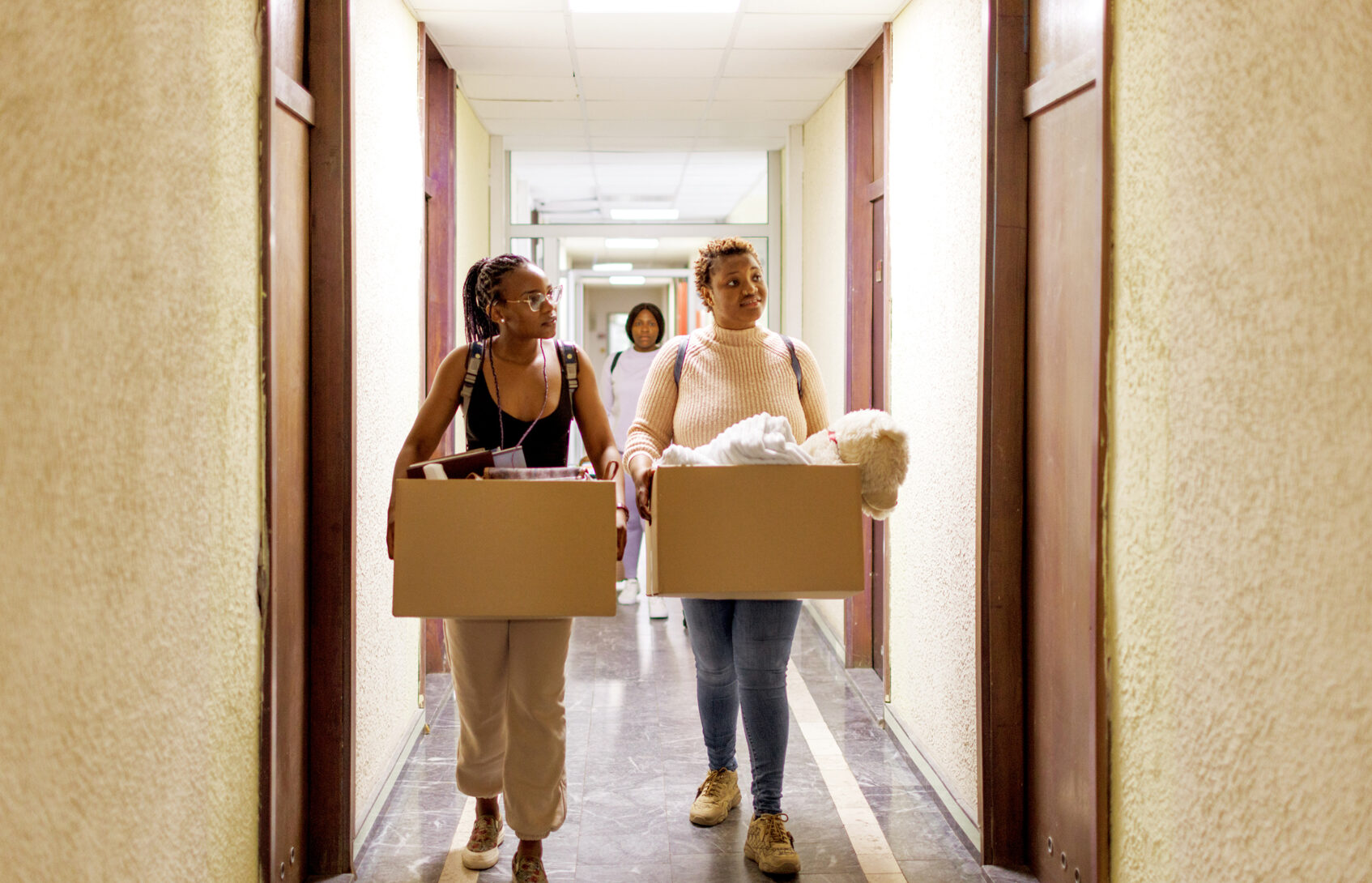 Two young women carrying boxes walking down the hallway of a college dorm
