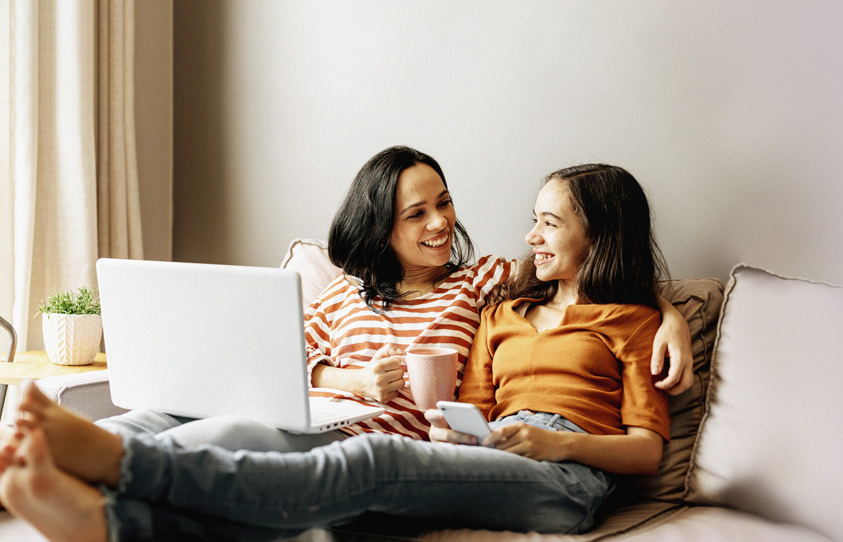 A mom and daughter sitting on a couch together smiling at each other, holding a laptop and a cell phone