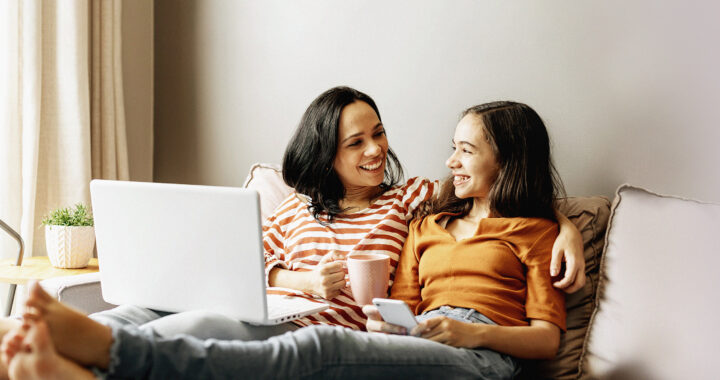 A mom and daughter sitting on a couch together smiling at each other, holding a laptop and a cell phone