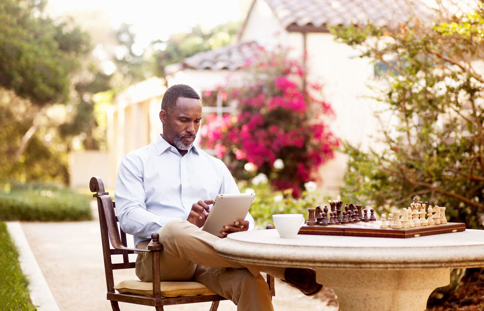 A man views a tablet while seated outside near a chess game.