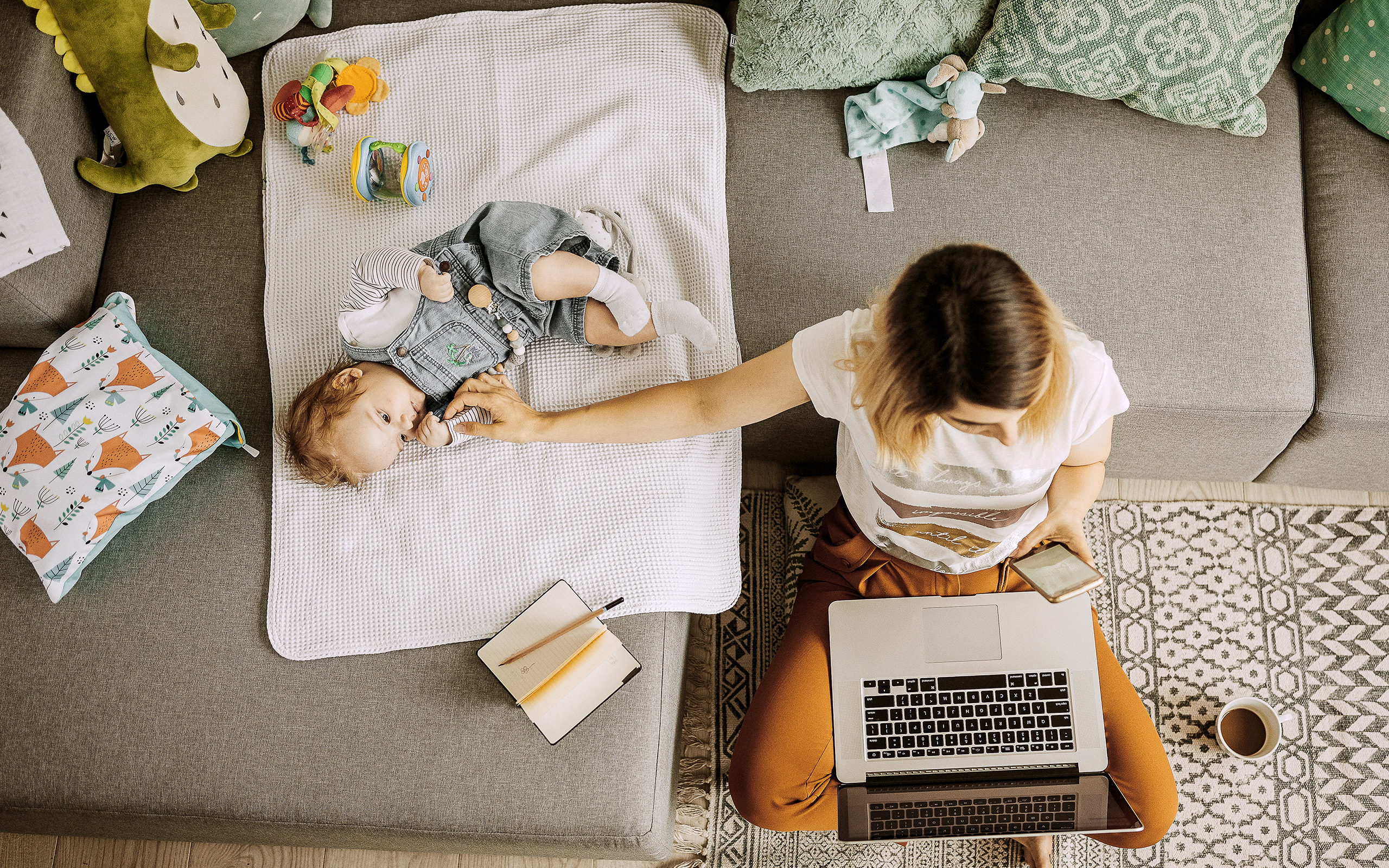 A mom sits on the floor with a laptop and a phone while holding one hand on her baby on the couch.