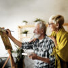 An older man smiling and painting with an older woman smiling and standing behind him.