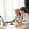 A mother and her adult daughter sit at a desk looking at a computer screen together.