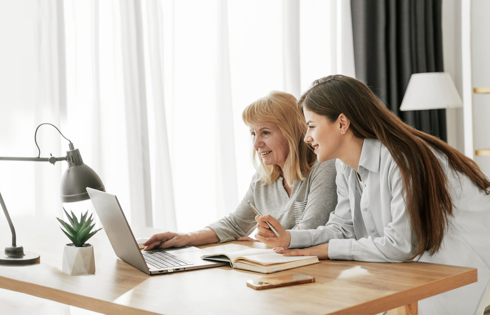 A mother and her adult daughter sit at a desk looking at a computer screen together.