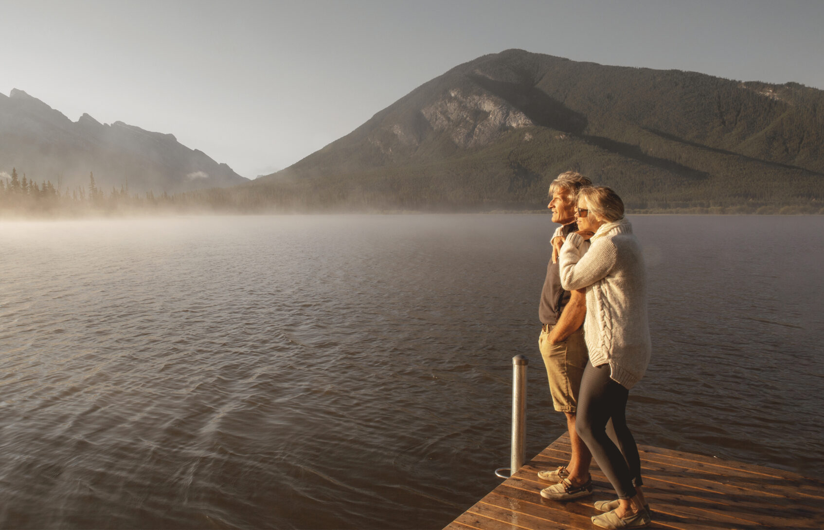 Couple standing on dock looks out over lake near mountains.