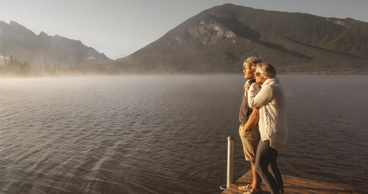 Couple standing on dock looks out over lake near mountains.